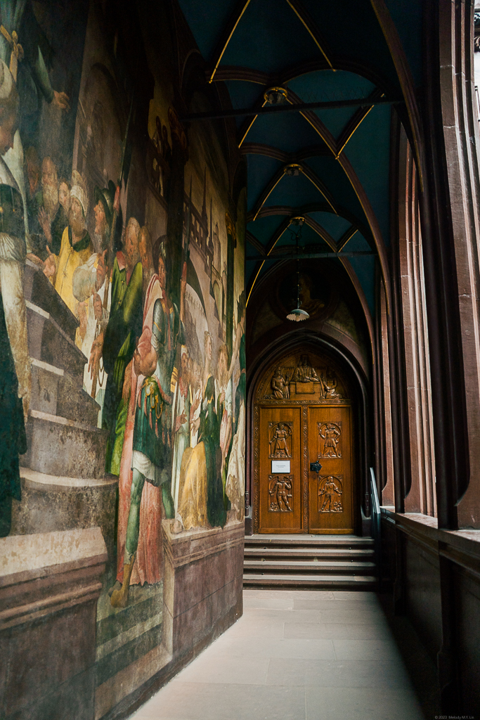 A carved, wooden door at the end of arched hallways; frescoes on the left wall