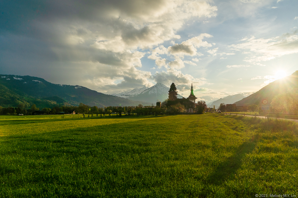 A small church across the field with the sun setting and hte mountains on the horizon